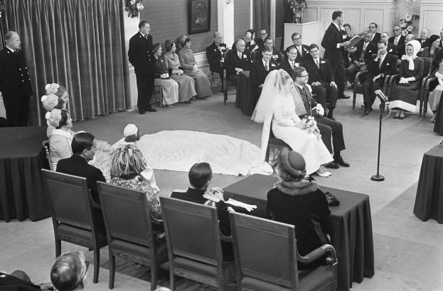 Schwarzes und weißes Foto einer Hochzeitszeremonie mit sitzenden und stehenden Menschen, einem Mann mit Mikrofon und einer Frau mit Blumenstrauß.