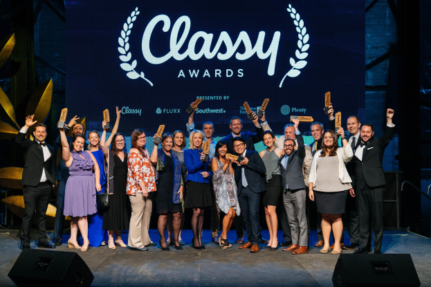 Gruppe von Menschen auf einer Bühne, die Auszeichnungen halten, mit Lautsprechern darunter und einem Bildschirm, der 'Classy Awards 2019' anzeigt.