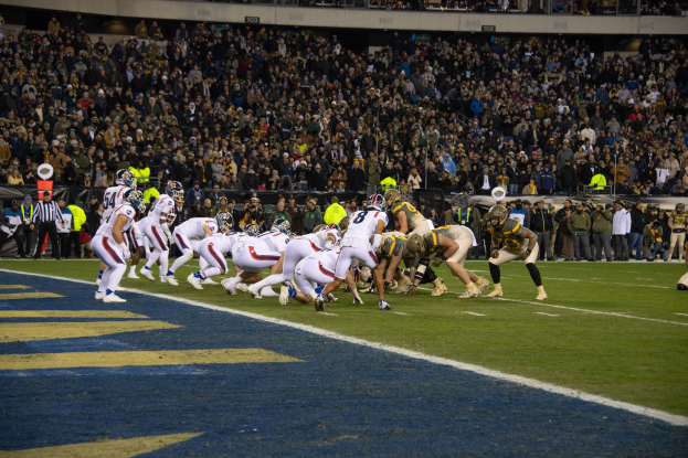 Ein Fußballspiel im Gange mit Spielern in Sportbekleidung und Helmen auf dem Boden, aus der Perspektive eines Stadionplatzes mit Zuschauern und Werbetafeln im Hintergrund.