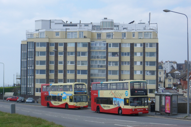 Zwei Doppeldeckerbusse fahren eine Straße mit hohen Gebäuden, Straßenlaternen, fahrenden Autos, Gras, Bäumen und einem klaren blauen Himmel im Hintergrund.