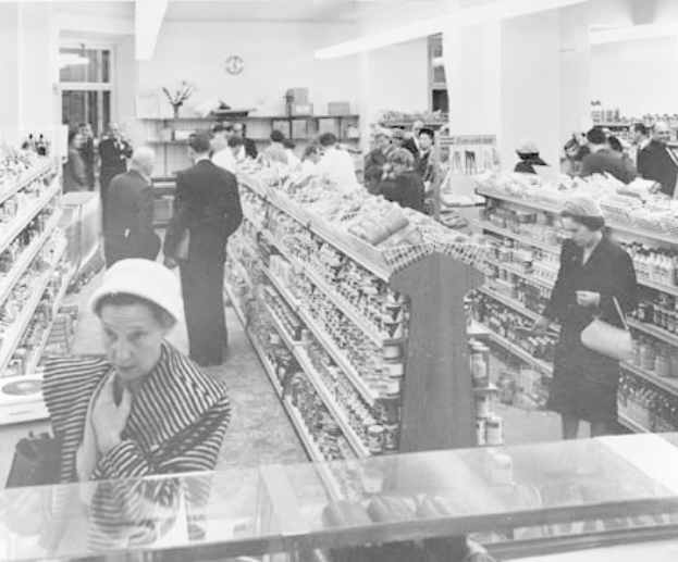 Schwarzes und weißes Foto von Menschen beim Einkaufen in einem Supermarkt mit Lebensmittelregalen, einige halten Taschen, eine Uhr an der Wand und Deckenbeleuchtung.