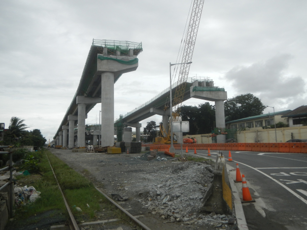 Baustelle mit einer Brücke im Hintergrund, Straße mit Verkehrskegeln markiert, Bahngeleise auf der linken Seite, verstreute Steine und Gras, Bäume und Gebäude auf beiden Seiten der Straße und ein bewölkter Himmel.