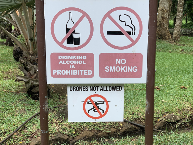 Schild am Straßenrand mit dem Text "Alkoholtrinken ist Verboten" und "Kein Rauchen" vor dem Hintergrund von Bäumen und Gras.
