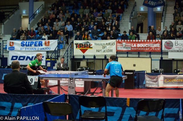 Indoor-Tischtennis-Match mit zwei Spielern, einigen Zuschauern im Stehen, sitzenden Zuschauern, leeren Stühlen und Werbeplakaten in einer Stadionatmosphäre.