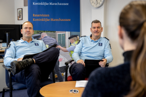 Zwei Polizeibeamte sitzen an einem Tisch mit Papieren und Gegenständen und sprechen mit einer Frau, im Hintergrund eine Wand mit einem Banner, einer Uhr und Fotorahmen, sowie ein weiterer Tisch links mit einem Monitor und Zubehör.