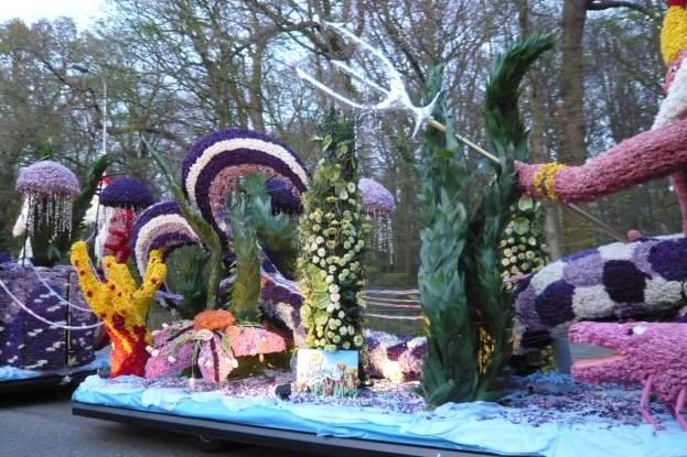 Ein Paradewagen, geschmückt mit einer Vielzahl bunter Blumen wie Rosen, Gänseblümchen und Lilien, umgeben von Bäumen unter einem klaren blauen Himmel.