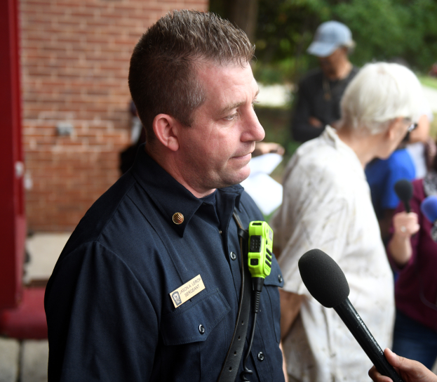 Ein Mann in Polizeiuniform spricht vor einer Backsteinwand mit Bäumen im Hintergrund zu Reportern, hält ein Mikrofon und ist von mehreren anderen Menschen umgeben, von denen einige ebenfalls Mikrofone halten.