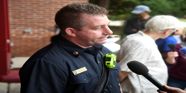 Ein Mann in Polizeiuniform spricht vor einer Backsteinwand mit Bäumen im Hintergrund zu Reportern, hält ein Mikrofon und ist von mehreren anderen Menschen umgeben, von denen einige ebenfalls Mikrofone halten.