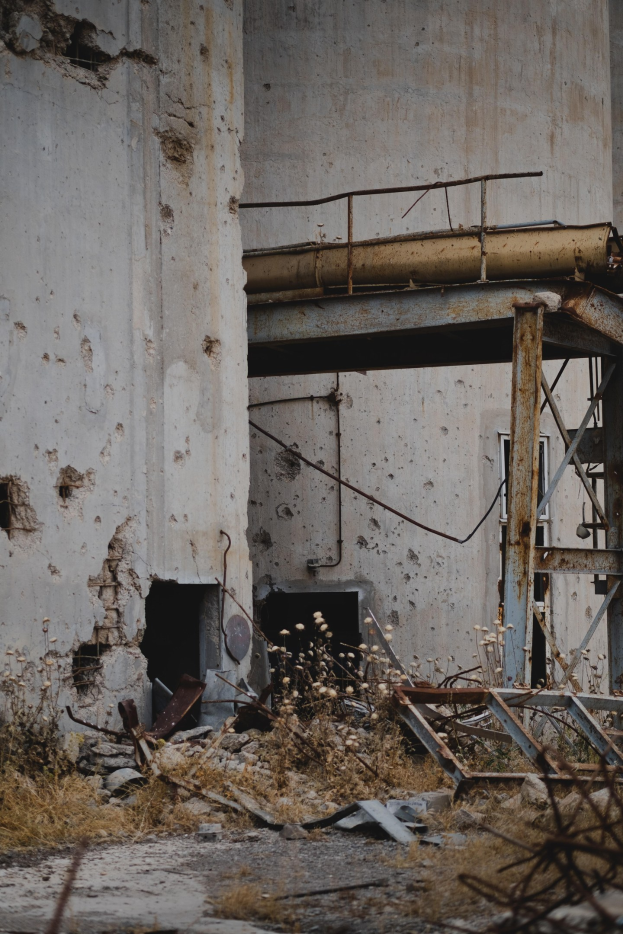 Altes Gebäude mit bröckelnden Wänden, kaputten Fenstern und einem zentralen Metallgerüst, umgeben von Pflanzen, Eisenstangen und Schutt.
