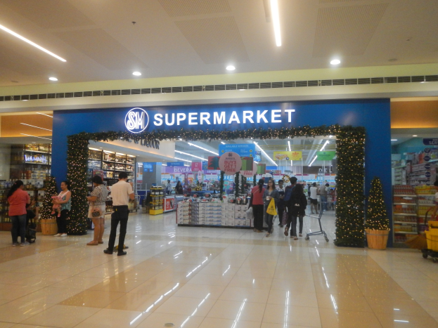 Gruppe von Menschen vor einem Supermarkt mit Einkaufstägern, Taschen, einem Einkaufswagen, Topfpflanze, Regalen, Schildern und Deckenleuchten.