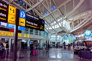 Ein Flughafen-Terminal-Inneneinrichtung mit Menschen, die gehen, einige tragen Taschen, und Pfosten mit Bändern und Texttafeln; Sofas und Tische mit Gegenständen auf der rechten Seite und ein Glaswand-Gebäude mit Deckeneinbauleuchten im Hintergrund.