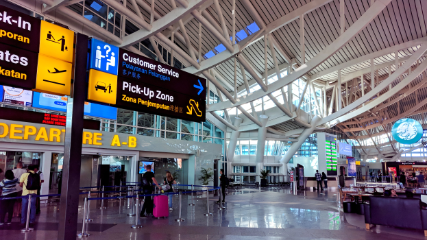 Ein Flughafen-Terminal-Inneneinrichtung mit Menschen, die gehen, einige tragen Taschen, und Pfosten mit Bändern und Texttafeln; Sofas und Tische mit Gegenständen auf der rechten Seite und ein Glaswand-Gebäude mit Deckeneinbauleuchten im Hintergrund.