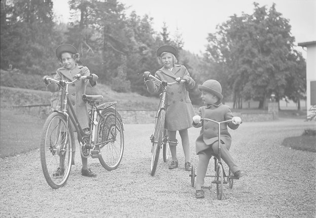 Drei Kinder in Mänteln und Mützen fahren Fahrräder in einem Park mit Bäumen und einem Haus im Hintergrund, aufgenommen in Schwarz-Weiß.
