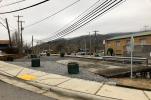 Eine Stadtstraße mit Gebäuden, Fahrzeugen, Bäumen und einem bewölkten Himmel, mit einem Gehweg, Mülltonnen, einer Straßenlaterne, Strommasten mit Drähten und einem Müllcontainer.