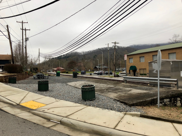 Eine Stadtstraße mit Gebäuden, Fahrzeugen, Bäumen und einem bewölkten Himmel, mit einem Gehweg, Mülltonnen, einer Straßenlaterne, Strommasten mit Drähten und einem Müllcontainer.