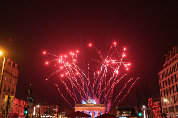 Eine belebte Stadtstraße mit Gebäuden, Bäumen, Laternen, Verkehrszeichen, Zeltplanen und Menschen, vor einem Nachthimmel mit farbenfrohen Feuerwerken während einer Silvesterfeier in Berlin.