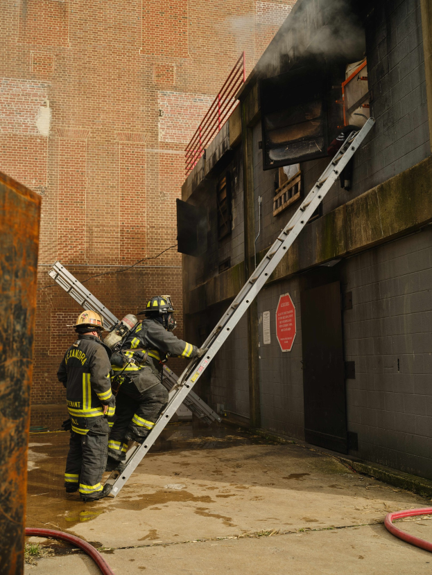 Feuerwehrleute in Helmen klettern an einer Leiter zu einem raucherfüllten Gebäude hinauf, mit einem Metallobjekt links und Rohren auf dem Boden darunter.