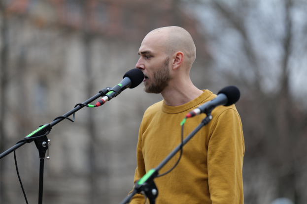 Ein kahlkopfiger Mann in einem gelben T-Shirt singt in ein Mikrofon mit einem unscharfen Hintergrund.
