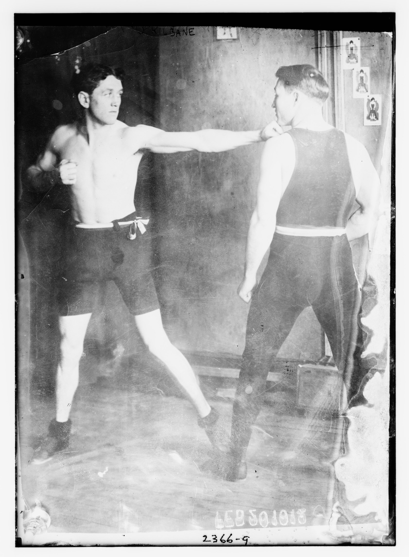 Schwarzes und weißes Foto von zwei Boxern in einem Ring mit Postern an der Hintergrundwand und Text unten.