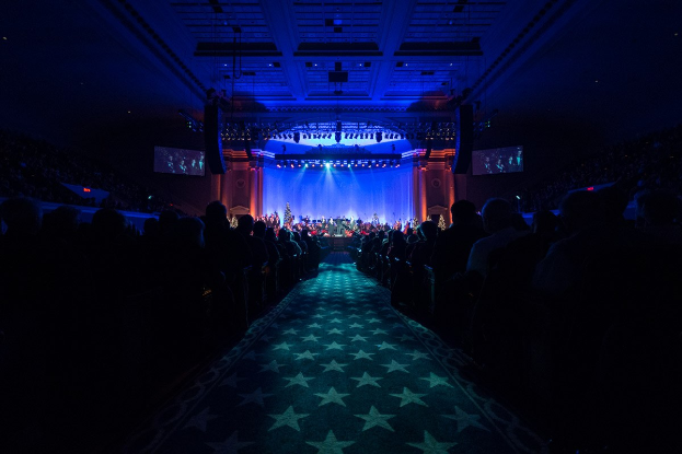 Ein großer Zuschauerraum mit Sitzplätzen, eine Bühne mit Musikern und Deckenleuchten und Fernseher, was auf eine Konzerthalle für die Nationale Convention 2019 hinweist.