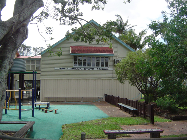 Ein Schulgebäude mit einem Spielplatz davor, umgeben von einem Zaun, Bänken, Gras, Pflanzen, Bäumen und anderen Gegenständen unter einem bewölkten Himmel.