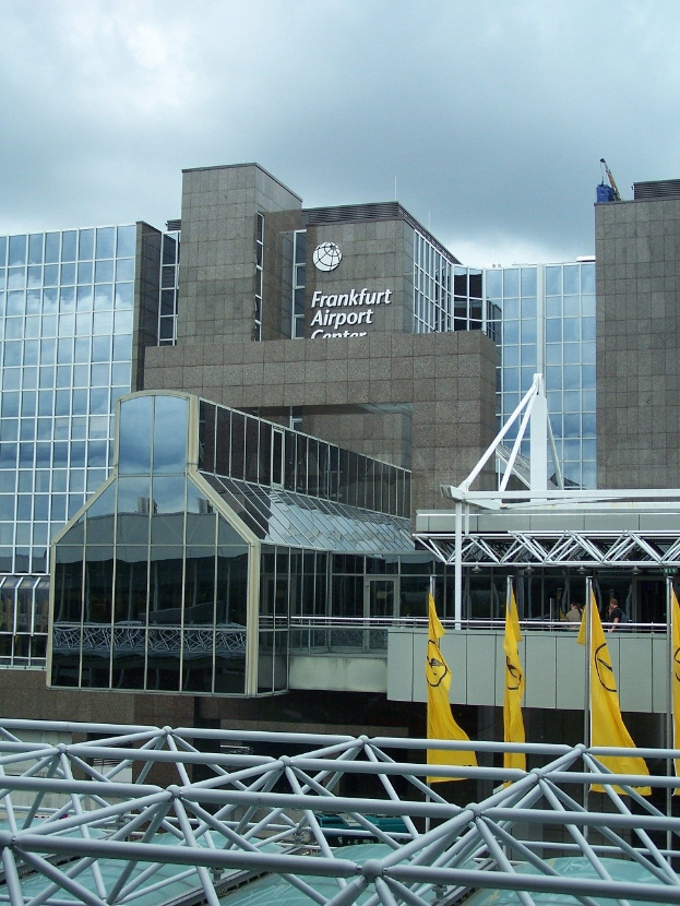 Frankfurt Airport in Frankfurt, Deutschland, mit einem großen Glasgebäude mit Text, umgeben von gelben Flaggen und Eisenstangen, unter einem Himmel mit weißen Wolken.