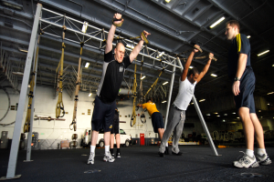 Eine Gruppe von Männern, die Klimmzüge in einem Fitnessstudio machen, wobei ein Mann in der Mitte Fußbekleidung trägt und von Metallstangen, Rahmen, einer Wand, einer Decke mit Deckenleuchten und verschiedenen Gegenständen auf der Oberfläche umgeben ist.