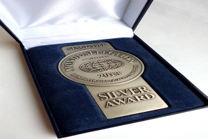 Eine silbergerahmte Auszeichnung mit eingravierter Inschrift wird in einer blauen Box auf einer weißen Oberfläche präsentiert.