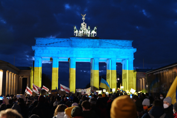 Menschenmenge mit Fahnen und Schildern vor dem Brandenburger Tor in Berlin, mit einem Banner auf der rechten Seite.