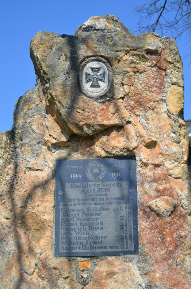 Mahnmal mit einer Tafel vor einem Gebäude, Baum im Hintergrund und Himmel darüber.