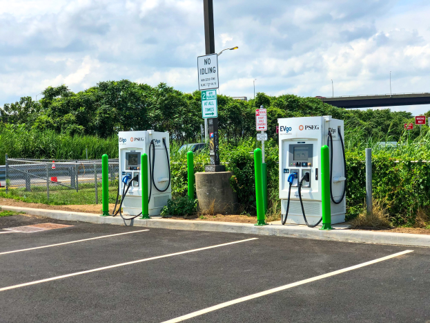 Elektrofahrzeug-Ladestation auf einem Parkplatz mit umgebender Infrastruktur, Vegetation und einer ├Übrückung im Hintergrund bei bew├Âlktem Himmel.