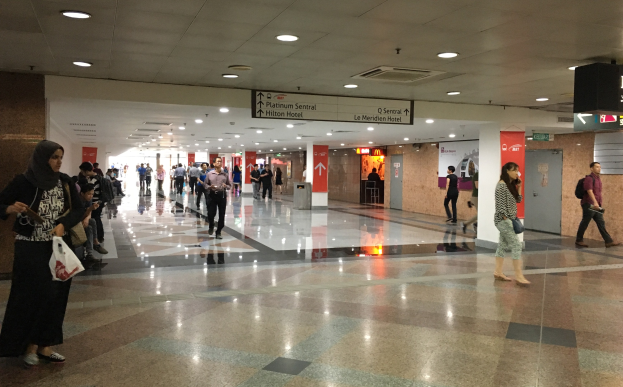 Menschen mit Gepäck durch eine Flughafenhalle gehen, Texttafeln an den Wänden, Deckenleuchten und ein Müllbehälter im Hintergrund; eine Frau in Schuhen ist zu sehen.