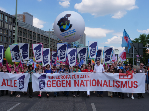 Eine Gruppe von Menschen geht eine Straße entlang und hält ein Transparent mit der Aufschrift "Ein Europa für alle gegen Nationalismus" und Fahnen, mit Gebäuden, Bäumen und einem bewölkten Himmel im Hintergrund.