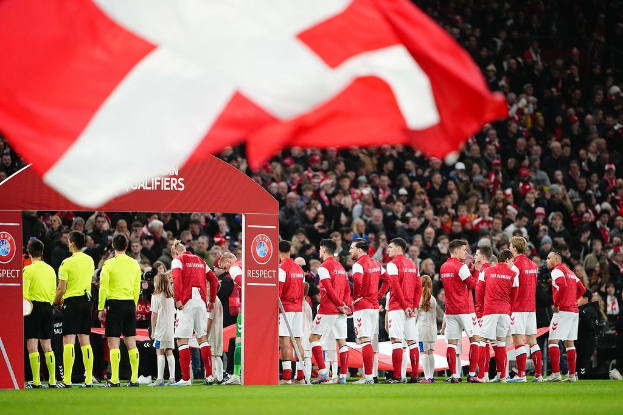 Eine Gruppe von Menschen auf einem Fußballfeld mit einer roten und weißen Flagge im Vordergrund, einem Bogen mit der Aufschrift "Bayern München vs Bayern München Wetten & Vorschau" im Hintergrund und einer großen Menschenmenge im Stadion.