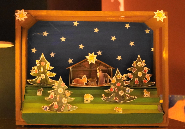 Eine Krippe des Heiligen Abends in einer Holzkiste auf einem Tisch, mit einer Lampe links und einer Wand im Hintergrund, mit dekorativen Weihnachtsbäumen, Tieren und einem Haus.