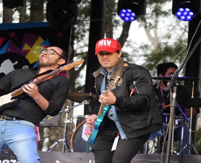 Zwei Männer auf der Bühne mit Gitarren spielend, mit einem Schlagzeuger dahinter, Mikrofone rundherum, unter Bäumen vor einem klaren blauen Himmel.