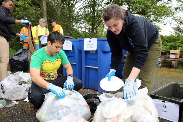 Eine Gruppe von Menschen, die Müll in einem Park sammeln, mit einem Mann und einer Frau in Handschuhen, die Teller halten, umgeben von Plastikmüll, einem Mülleimer, einer Bank, Bäumen und einem klaren blauen Himmel.