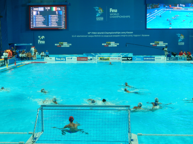 Eine Gruppe von Menschen spielt Wasserball in einem großen Pool mit einem Netz in der Mitte, Zuschauern auf der linken Seite und einem Bildschirm mit Text und Logos im Hintergrund bei den 2016 FINA-Weltmeisterschaften in Kazan.