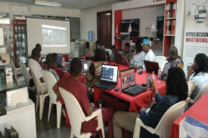 Gruppe von Menschen an einem Tisch mit Laptops und anderen Gegenständen während eines digitalen Marketing-Workshops, mit einem Banner, einem Display und Deckenleuchten im Hintergrund.