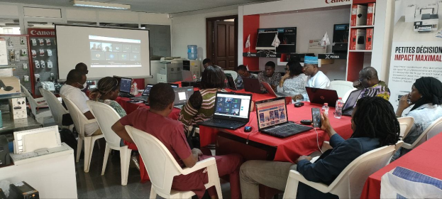 Gruppe von Menschen an einem Tisch mit Laptops und anderen Gegenständen während eines digitalen Marketing-Workshops, mit einem Banner, einem Display und Deckenleuchten im Hintergrund.