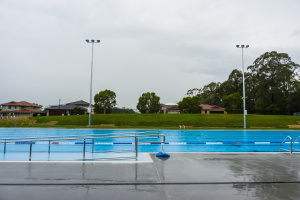 Große rechteckige Schwimmbad in einem Park, umgeben von Zäunen und Pfählen mit Oberlichtern, umgeben von Bäumen, mit Häusern und einem klaren blauen Himmel im Hintergrund.