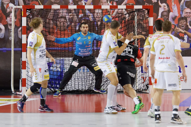 Mannergruppe spielt Handball auf einem Feld mit einem Ball im Spiel, in einer Halle mit einem Netz und einer Fahne im Hintergrund.