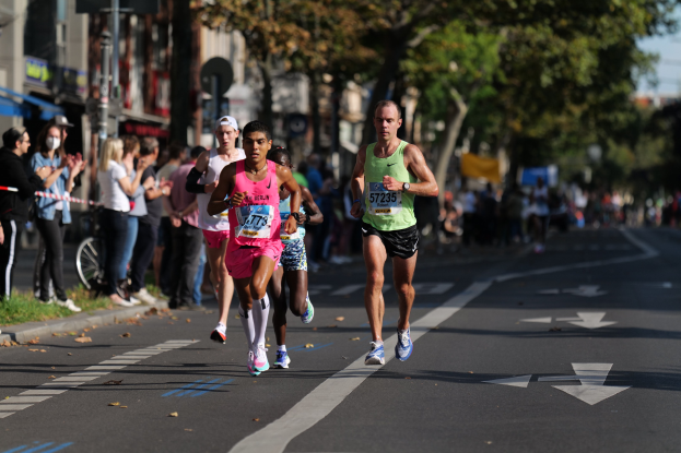 Eine Gruppe von Menschen, die bei einem Marathon auf einer Stadtstraße laufen, mit Zuschauern auf der linken Seite, unscharfen Gebäuden, Bäumen und einem Fahrrad im Hintergrund.