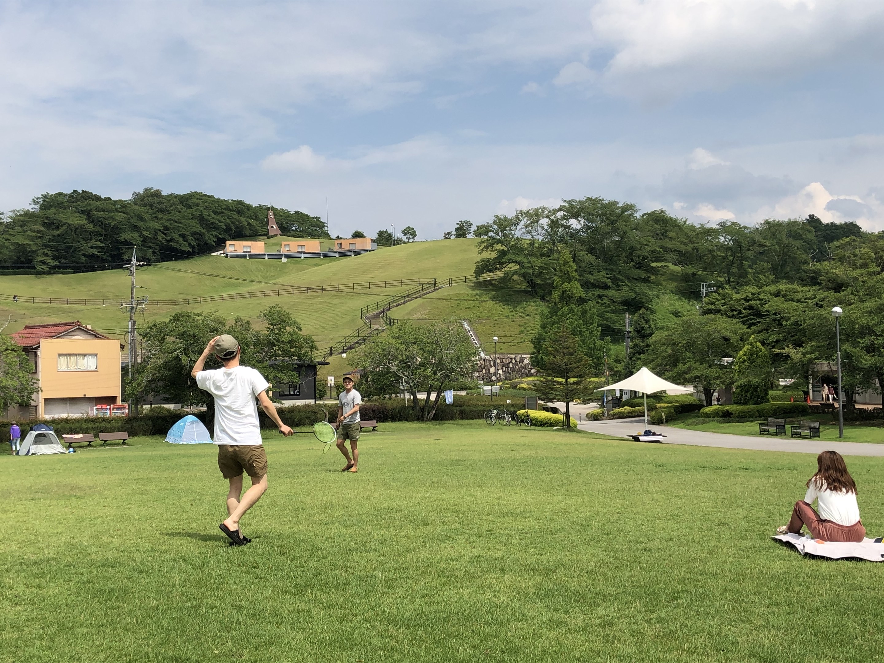Gruppe von Menschen, die Badminton in einem Park spielt, mit einem Mann, der einen Schläger hält und auf Gras in der Nähe von Zelten und Gebäuden sitzt, mit Hügeln und bewölktem Himmel im Hintergrund.