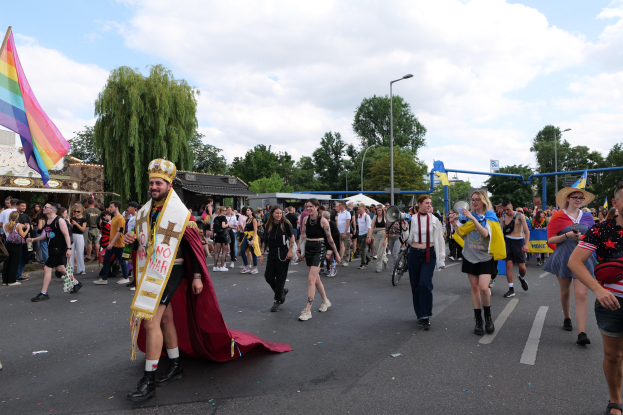 Eine Gruppe von Menschen marschiert bei der Christopher Street Day Parade 2018 mit einer Regenbogenflagge und Musikinstrumenten, während im Hintergrund Laternenmasten, Bäume und Hütten zu sehen sind.