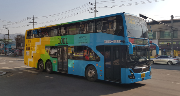 Ein blauer und gelber Doppeldeckerbus fährt auf einer Straße mit anderen Fahrzeugen, umgeben von Gebäuden, Bäumen, Strommasten und einem klaren Himmel.