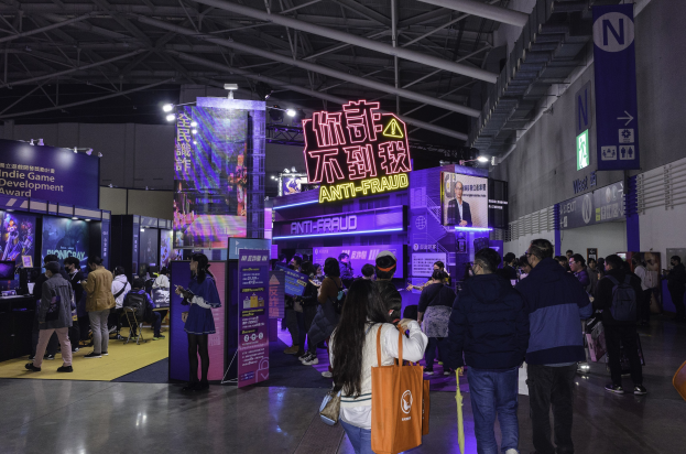 Eine große Gruppe von Menschen steht vor einem "Indie Game Development Award 2019"-Stand auf einer Messe, wobei einige Taschen und Stände mit Bildschirmen und Text im Hintergrund halten.