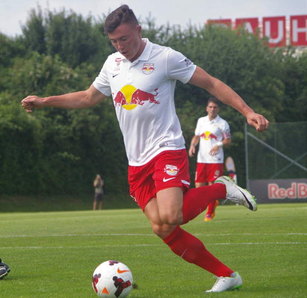 Ein professioneller Fußballspieler kickt einen Ball auf einem grünen Feld mit Bäumen und einem klaren blauen Himmel im Hintergrund, wobei ein "RB Leipzig"-Schild sichtbar ist.