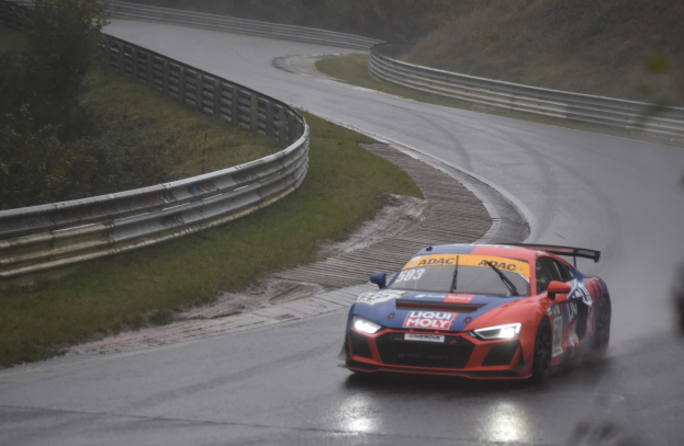 Audi R8 LMS GT3 Rennwagen auf der Nürburgring 24 Stunden Strecke, umgeben von grünem Gras und Bäumen mit einem Geländer im Hintergrund.