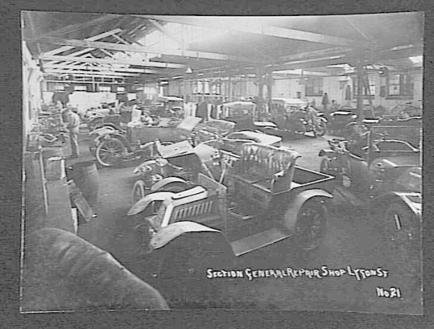 Schwarze und weiße Fotografie einer frühen 20. Jahrhundert-Autowerkstatt in Lyon, Frankreich, die Fahrzeuge, Arbeiter, Werkzeuge und Decken Details mit Text am Boden zeigt.